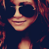 A close-up of a person with long wavy brown hair wearing dark aviator sunglasses and a light-colored top, cropped to show only the upper face and shoulders.