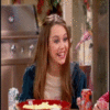 A person with long brown hair wearing a dark blue jacket over a light top, seated at a table with a red bowl of food and a red can, in a kitchen setting with stainless steel appliances.