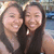Two women with long dark hair wearing sleeveless tops pose closely together, smiling at the camera in a bright outdoor setting.