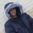 Pixelated figure in dark hooded jacket with light-colored hood, standing against light background with faint beige patch on right.