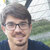 Pixelated portrait of a person with short dark hair, glasses, and a mustache, wearing a dark blue shirt against a blurred outdoor background.