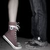 Two people's lower legs and feet stand on a textured surface; one wears a maroon high-top sneaker with white laces and a white stripe, the other dark jeans with a silver button.