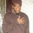 Pixelated figure in brown turtleneck sweater with hand near chest, standing against light background.