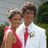 A static image shows two people in formal attire standing close together outdoors, with greenery and a light-colored wall behind them.
