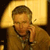 Pixelated image of a person with light brown hair wearing a brown shirt, holding a phone to their ear against a yellow-lit background.