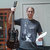Pixelated image of a person holding a long object, standing next to a bookshelf with a framed picture on the wall.