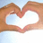 Two hands form a heart shape against a light blue sky background with white clouds.