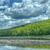 Pixelated landscape with green hills, blue sky, and white clouds over a reflective water surface.