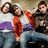 Three people sit together in a , two wearing white and purple striped shirts with orange accents, one in brown jacket; the rightmost person's arm moves from raised to lowered position