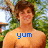 Pixelated image of a shirtless person with brown hair standing outdoors, green foliage background, blue text 'yum' overlaid on chest.