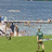 Pixelated image of a person in green shirt and brown shorts on grassy field with water and structures in background, overlaid with pink text on blue background.