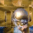 Pixelated static image of a person with light-colored hair and glasses, wearing a white shirt against a brown wooden background.