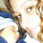 Pixelated image of a person with light skin and brown hair wearing blue clothing, holding an object near their face against a bright background.