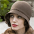 A pixelated close-up of a person wearing a light brown cloche hat, dark brown hair, and a brown coat with a fur collar, set against a blurred green and white background.