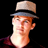 Pixelated static image of a person wearing a light-colored fedora-style hat with dark band, red hair, and visible neck against black background.