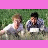 Two figures in white and dark clothing sit on grassy ground against a green background with pink borders.