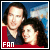 Two people with dark hair pose closely together against a blue sky background, one wearing red clothing and the other in dark attire.