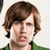 Pixelated close-up of a person with brown hair, light skin, and a green garment visible at the bottom edge, set against a light background.