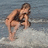 Pixelated image of a person in a crouched pose on a beach, wearing dark swimwear against a blue-gray water background.