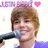 A person with light brown hair holds a dark blue microphone near their mouth, wearing a purple shirt against a white background with repeated text and pink hearts.
