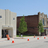 A pixelated outdoor scene shows a light-colored building with a dark entrance, green trees, and orange traffic cones on a paved surface under a clear blue sky.