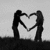 Two silhouetted figures stand side-by-side, their arms raised to form a heart shape with their hands against a light gray background.