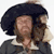 Pixelated image of a person with dark curly hair wearing a dark jacket over a light collared shirt, holding a small animal against their chest.