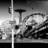 Black-and-white image of a street scene with a tall tower, a lamppost, and a large vehicle on a road.