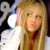 A person with long blonde hair and bangs wears a white jacket with a blue button, holding a hand near their head against a dark background with blurred lights.