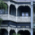 A pixelated static image of a multi-story building with arched windows and a balcony, set against a blurred background with greenery.