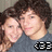 Two people with dark curly hair pose closely together, one wearing a dark blue shirt with white text on the bottom right.