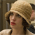 A pixelated close-up of a person wearing a light brown wide-brimmed hat with a dark band, dark curly hair, and red lips, set against a blurred background.