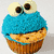 A cupcake with blue frosting shaped like a character's face, featuring two large round eyes with brown pupils and white sclera, on a light background.