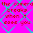 A static image with a bright pink background overlaid by cyan diamond-shaped lines, displaying repeated red text that reads "the camera breaks when it sees you" in pixelated font.