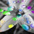 Pixelated static image of a stylized figure with gray and white tones, green accents on limbs, blue highlights on shoulders, yellow circular elements near the base, purple and cyan scattered pixels in background.