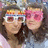 Two people with long brown hair pose closely together; one wears white-framed glasses, the other pink heart-shaped sunglasses against a blurred background.