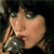 A pixelated close-up of a person holding a microphone near their mouth, with dark hair and a dark background.