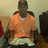 Pixelated image of a person in an orange shirt and light pants seated with arms crossed against a muted background.