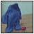 Pixelated blue figure in a hooded coat stands on sandy ground with a red object near its feet against a light blue sky.