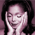 Pixelated image of a person with dark hair holding both hands near their face, closed eyes, and a slight smile against a purple background.