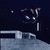 Pixelated dark blue background with a small light-colored circular object on the left, and a stylized figure holding a long vertical object in the center-right.