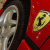 Red background with repeated yellow Ferrari prancing horse logo and silver car wheel, text "FERRARI" in white pixelated font across the bottom.