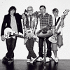 Black-and-white photograph of a band with two identical groups of five people holding guitars and a drum set, with pink text below reading "wich do yew prefer???".