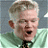 Pixelated close-up of a person with light brown hair, wearing a green shirt and dark tie, mouth open in mid-expression against a blurred background.