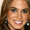 A close-up portrait of a person with light brown hair, wearing dark eyeliner, red lipstick, and dangling earrings with white and dark beads.