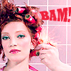 A woman with red hair in curlers holds a red spray bottle, standing against a pink grid background with red text reading "BAM!".
