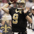 A football player in a black jersey with gold number 9 and fleur-de-lis logo, helmet on, arms raised, standing on a field.