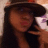 A pixelated close-up of a person with long dark hair wearing a dark hat, holding a white object near their face.