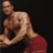 Static pixelated image of two shirtless men in close physical contact against a dark background, with one man embracing the other from behind.