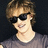 Pixelated close-up of a person with light brown hair wearing dark sunglasses and a gray shirt against a black background.